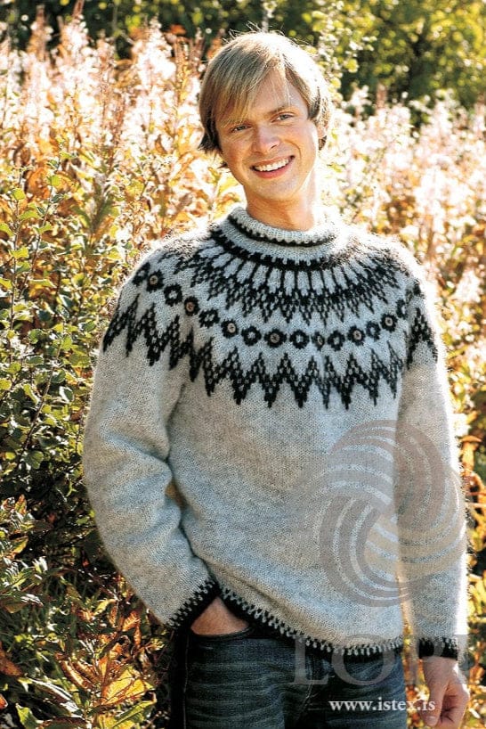 Smiling young man with fair hair in front of shrubs and tall flowering plants in autumn colours, wearing Hringur in the pattern’s own colours — a light ash heather pullover with a round yoke of black heather and chocolate: a band of large zigzags, a row of small dark rings with a light wooden button in every other one, a band of dark points and a dark band near the neck, a folded-down collar, and dark speckled edges at the hem and cuffs. The blue jeans are not part of the pattern