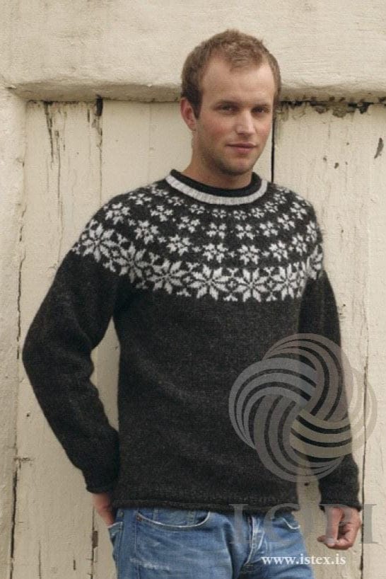 Young man with short reddish-blond hair in front of weathered white-painted wooden boards, wearing Fönn in the pattern’s own colours — a black heather sweater with a yoke of ash heather eight-pointed stars, large at the base and smaller above, an ash heather ribbed neckband with a dark rolled edge, and rolled edges at the cuffs and hem. The blue jeans are not part of the pattern. The Lopi logo and www.istex.is are printed on the photo