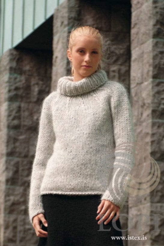 Young woman with blonde hair pulled back, standing in front of rough stone pillars, wearing Nú in the pattern’s light grey (ash heather) — a plain bulky sweater with a round yoke, the knitted-on ribbed collar folded over, and a rolled hem. The small stud earring and the black skirt are not part of the pattern. The Lopi logo and www.istex.is are printed on the photo