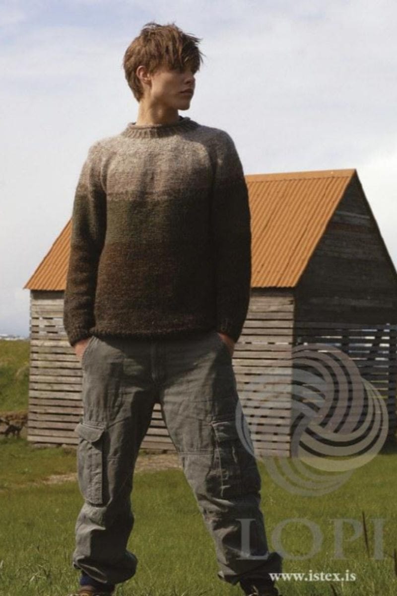 Young man with brown hair in front of a wooden shed with an orange corrugated roof, wearing the Moss version of Útjörð in the pattern’s own colours — a round-yoke sweater, darkest at the hem, with a band of spruce green across the chest and lighter beige towards the neck, and ribbed hem, cuffs and neck. The grey cargo trousers, the socks and the shoes are not part of the pattern
