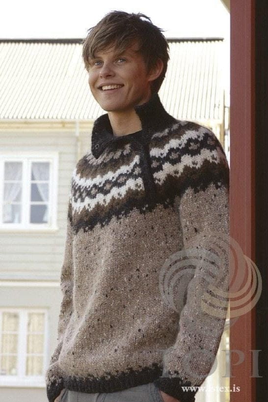 Smiling young man standing by a red post, a pale house with white windows behind him, in the brown Órói, shown in the pattern’s original colours (beige tweed is no longer made by Ístex) — a flecked beige tweed Álafosslopi sweater with black heather ribs, a round yoke in bands of black heather, white and acorn, and a high black heather collar over an opening to one side of the front. The grey trousers are not part of the pattern