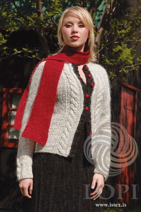 Young woman with blonde hair in front of a dark wooden house with red window and door frames, in Gréta, shown in the pattern’s own colours — an ash-grey Jöklalopi cardigan with a cable up each front, a short cable at the lower sleeve and dark grey ribbed front bands with red buttons. The red scarf and the long dark grey dress under the cardigan are not part of the pattern; the buttons are not included
