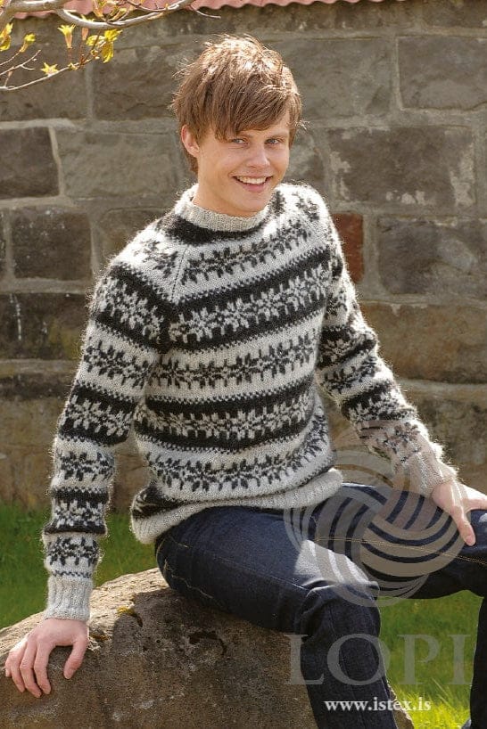 Smiling young man sitting on a large rock in front of a stone wall, in Nost, shown in the pattern’s own colours — an ash Léttlopi raglan sweater patterned all over with bands of black heather eight-petal roses on ash and ash roses on black heather, with ash ribs at the hem, cuffs and neck. The dark blue jeans are not part of the pattern