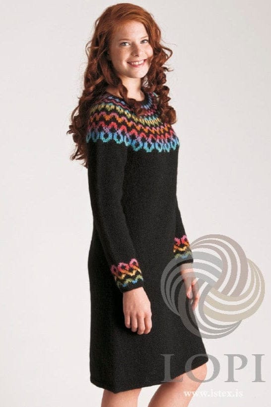 Smiling young woman with long curly red hair against a pale background, wearing the long Keðja in the pattern’s original eight-colour version — a knee-length black dress with a yoke of wavy bands in blue, lilac, red, orange, pink, green and yellow, a narrower band of pink, red, orange, yellow, green and blue above the cuffs, and a plain black hem