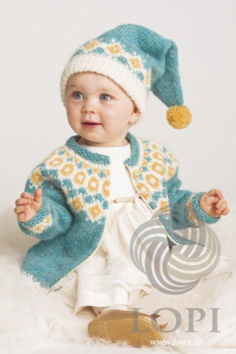 Baby sitting on a white fluffy rug, wearing Kría in the pattern’s bluegreen colours — an open glacier blue cardigan with a white yoke of sun yellow rings and diamonds and small glacier blue motifs, the same colours above the cuffs, a glacier blue neckband and a light zip, and the long pointed hat with a white folded brim, a band of yellow and blue on white, and a yellow pom-pom. The white dress with a ribbon bow and the light brown shoes are not part of the pattern, and the zip is not included