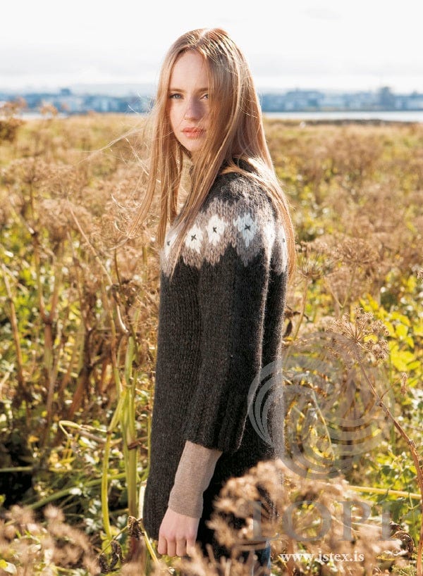 The same young woman turned to the side among the dry plants, in the same long black Rosa with chart B — the oatmeal band with embroidered white diamonds and a ¾ sleeve with its twisted-rib cuff. The beige long-sleeved top under the sleeve and the jeans just showing below the hem are not part of the pattern