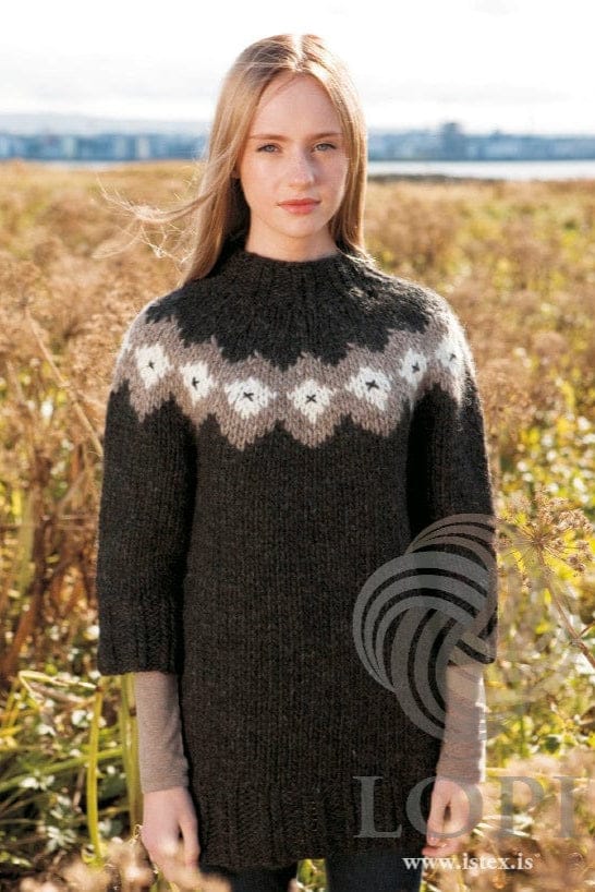 Young woman with long straight blonde hair standing among tall dry plants, water and a town far behind, wearing the long black Rosa with chart B in the pattern’s own colours — black heather with ¾ sleeves and a high twisted-rib neck, and a yoke band of oatmeal with white diamonds, each with a small black X. The beige long-sleeved top under the sleeves and the dark trousers below the hem are not part of the pattern