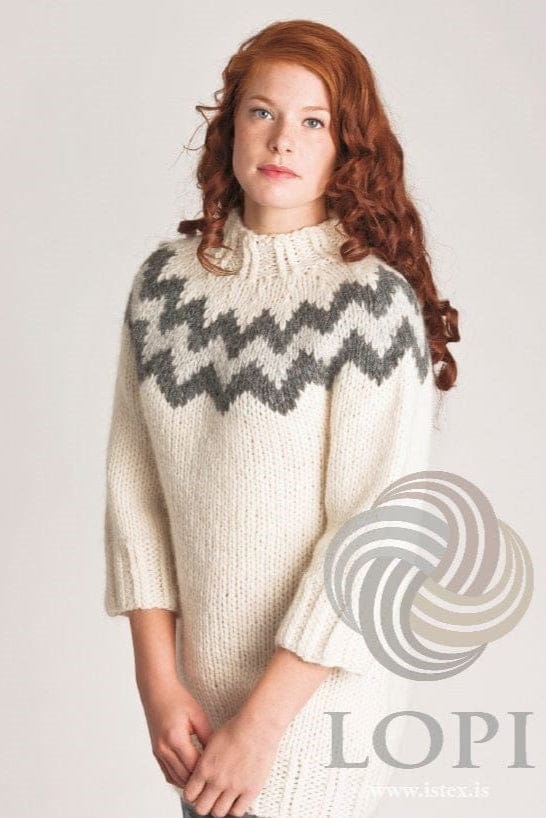 Young woman with long curly red hair against a pale grey background, wearing the long white Rosa with chart A in the pattern’s own colours — white with ¾ sleeves, twisted-rib cuffs and a high twisted-rib neck, and a yoke with a wide dark grey zigzag band and a lighter zigzag inside it. The jeans just showing below the hem are not part of the pattern