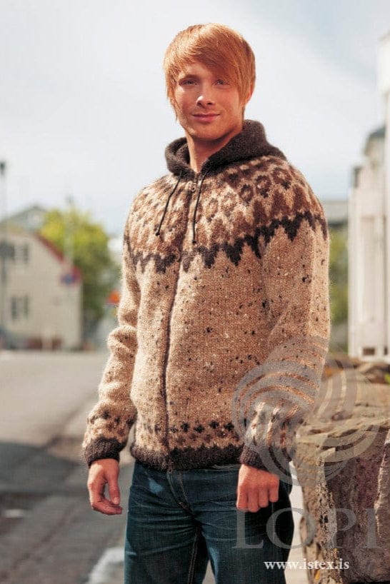 Young man on a town street in the brown Frost, shown in the pattern’s original colours (beige tweed is no longer made by Ístex) — a flecked beige tweed Álafosslopi zip cardigan with a dark brown hood, ribs and zigzag band at the base of the yoke, acorn-brown motifs above it, a border of small brown crosses and dots above the hem and cuffs, and a dark cord at the neck. The blue jeans are not part of the pattern; the zipper and cord are not included