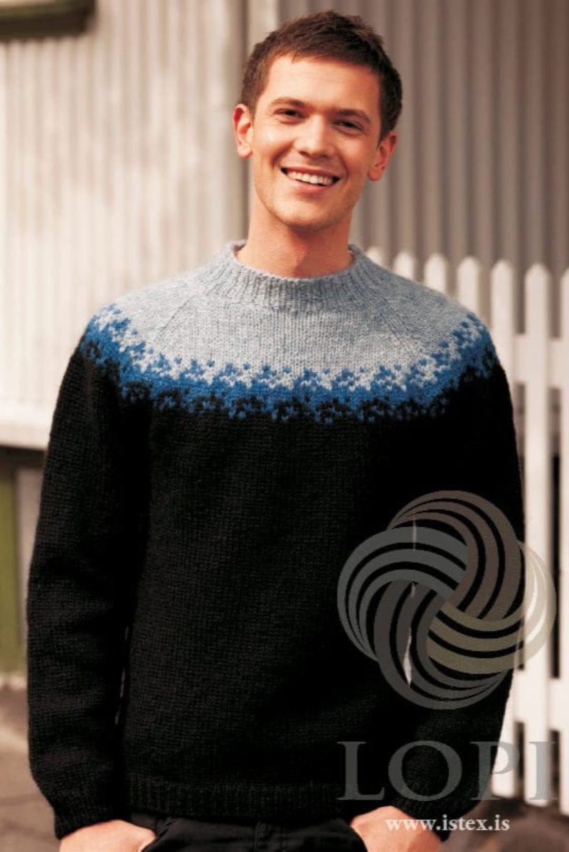 Young man by a white picket fence in the black-blue Sátt, shown in the pattern’s original colours (faded denim is no longer made) — a black Plötulopi sweater with an arctic blue jagged band and a light blue-grey upper yoke with raglan lines and neckband. The dark trousers are not part of the pattern