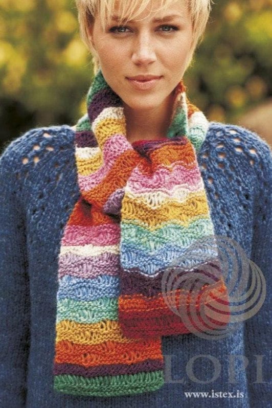 Young woman with short blonde hair wearing Alda wrapped round her neck, shown in the pattern’s original colours (orange, golden and lilac are no longer made) — a striped Léttlopi scarf in garnet, orange, violet, pink, golden yellow, pale grey, blues, lagoon and lilac, with long dropped stitches making wavy lines in each stripe. The blue sweater with eyelets on the shoulders is not part of the pattern