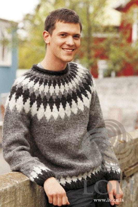Smiling young man with short dark hair leaning on a wall, a red house behind, wearing the grey Tíglar in the pattern’s own colours — grey with a yoke of black heather and ash zigzag bands, black heather rib and neck, and ash points above the hem. The light grey T-shirt at the neck and the dark trousers are not part of the pattern