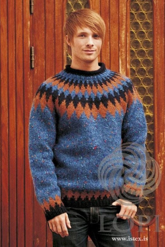 Young man with reddish fair hair in front of a wooden door, wearing the blue Tíglar in the pattern’s original colours — monet blue with coloured flecks, a yoke of amber and midnight blue zigzag bands, midnight blue rib and neck, and amber points above the hem and cuffs. The jeans are not part of the pattern