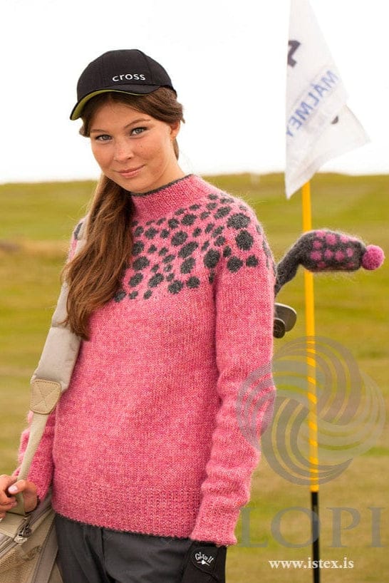 Young woman on a golf course in the pink Gola sweater, shown in the pattern’s own colours — sunset rose and blush with dark grey spots scattered over the yoke; behind her the dark grey golf club cover with pink spots and a pink pompom, also in the pattern. This photo is from Ístex’s photo set for the pattern, not the PDF. The cap, the golf bag and its strap, the glove, the trousers, the flag and the club under the cover are not part of the pattern