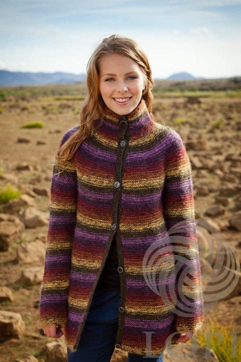Smiling young woman on a stony plain with hills behind in the colourful Katla, shown in the pattern’s original colours — bands of golden yellow, dark amber, jasper red, plum and hyacinth with darker marsh bands, the collar standing up and buttoned. The dark top under it and the blue jeans are not part of the pattern
