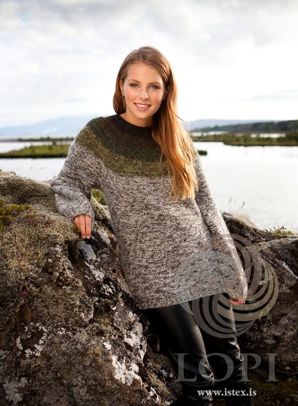 Smiling woman leaning on a mossy rock by a lake in the greentone Rangur, shown in the pattern’s own colours, purl side out — marled ivory beige below, darkening green bands in the yoke and a dark neckband. The black leather trousers are not part of the pattern