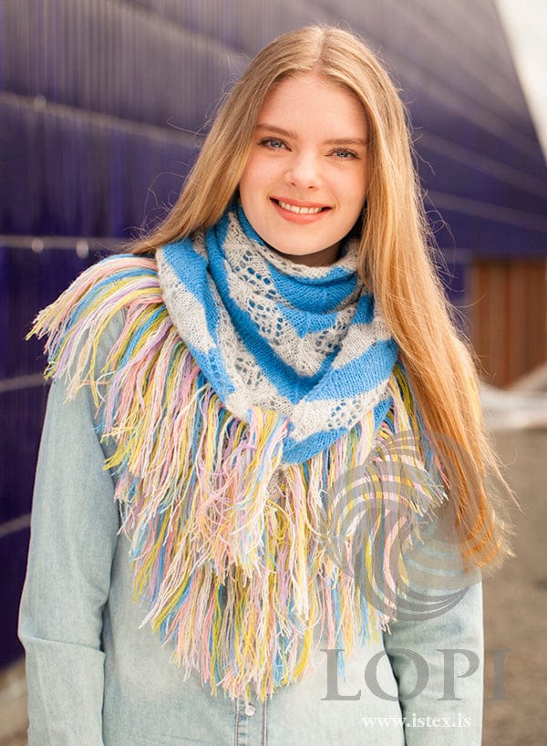 The same young woman, smiling, with Vængur wrapped round her neck like a scarf and the point and fringe hanging at the front, in the pattern’s own colours — sky blue and ash-heather lace stripes with the six-colour fringe. The light blue denim top is not part of the pattern