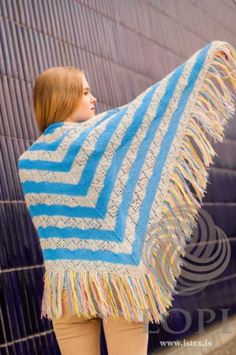 Young woman seen from behind against a wall of dark blue tiles, holding one arm out to spread Vængur, shown in the pattern’s own colours — bands of sky blue stocking stitch and ash-heather lace meeting in a V at the centre back, and a long six-colour fringe along both sloping edges. The beige trousers and the light blue top showing at her waist are not part of the pattern