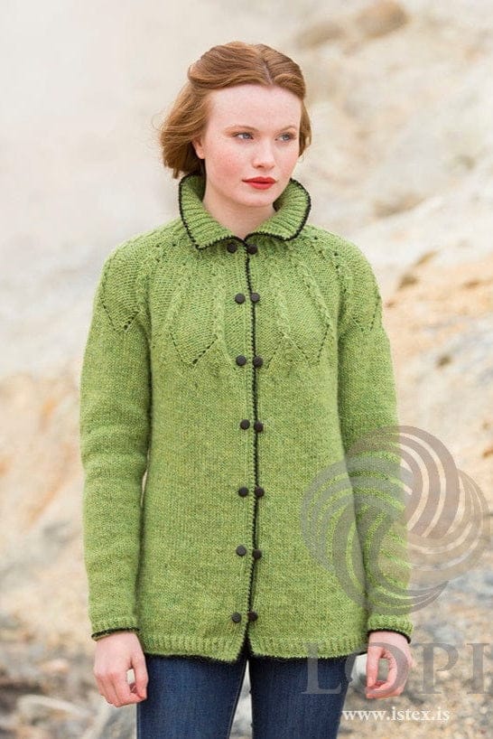 Young woman with red hair in front of pale sandy rocks in Vor, shown in the pattern’s own colours — a spring green Léttlopi cardigan with a fold-over collar, a textured yoke of purl panels outlined with eyelets, black crab-stitch edges down the fronts and round the collar, hem and cuffs, and two rows of round dark buttons. The blue jeans are not part of the pattern, and the buttons are not included