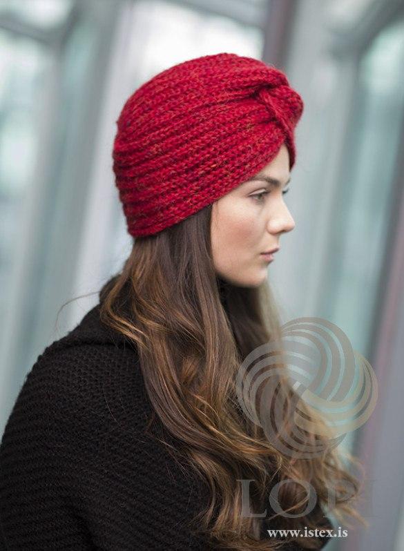 Woman in profile wearing Höttur in carmine red heather, the pattern’s own colour — a turban of brioche ribs running round the head, with the knot at the front. The black knitted top is not part of the pattern