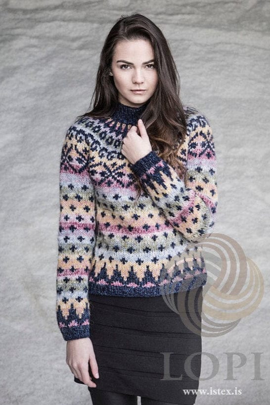 The Auðna sweater without the hat, against a pale grey wall, shown in the pattern’s original colours — navy stand-up collar and cuffs, blended colour bands with small navy crosses and a navy-patterned yoke. The black skirt and black tights are not part of the pattern