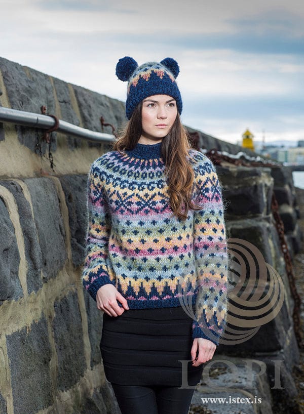 Young woman by a stone harbour wall in Auðna and Happ, shown in the pattern’s original colours (three of them are no longer made) — a Plötulopi sweater with navy rib, bands of lilac, pink, yellow, light blue and green dotted with small navy crosses and a yoke of navy motifs, and the matching hat with a deep navy rib and two navy pom-poms. The black skirt and black leggings are not part of the pattern