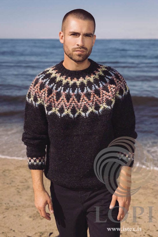 Bearded man on a sandy beach by the sea wearing Marbendill, shown in the pattern’s own colours — black, with a round yoke of sky blue, lime, crimson and orange around large black diamond shapes, and a small coloured band above each ribbed cuff. The black trousers are not part of the pattern