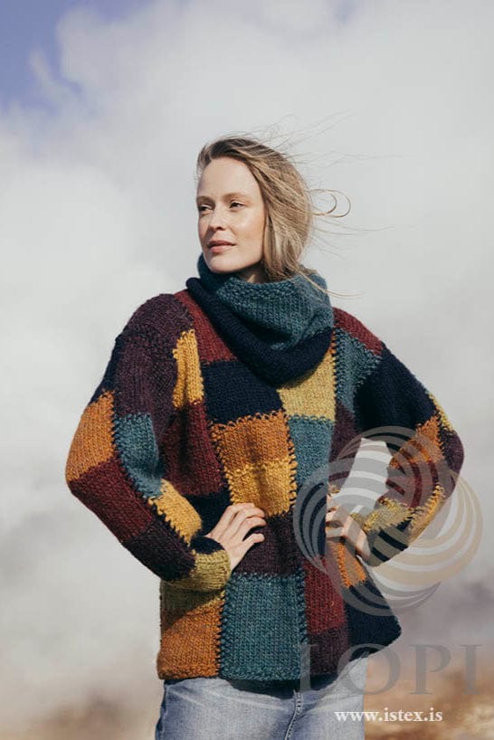 Woman with her hands on her hips in the colorful Brautir and its snood, shown in the pattern’s own colours — blocks of midnight blue, bordeaux, ruby red, golden, amber, chartreuse green and teal, and a snood in midnight blue and teal. The light jeans are not part of the pattern