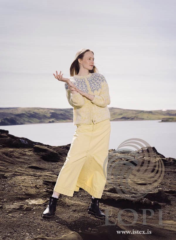 Woman on dark rocky ground above a lake, one hand raised, wearing Bláregn Open in the pattern’s own colours — a short cardigan in a yellow-and-white marl of white Plötulopi and yellow Einband, with clusters of small light blue flowers round the yoke, closed down the front. The long pale yellow skirt, the white socks, the black ankle boots and the bracelet are not part of the pattern