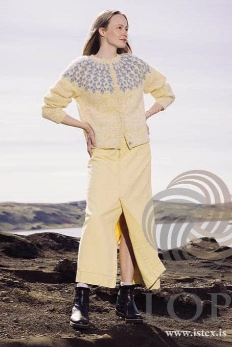 Bláregn Open worn with hands on hips, in the pattern’s own colours — the yellow-and-white marl body and sleeves, the light blue flower yoke and the folded front bands. The long pale yellow skirt with a front slit, the white socks, the black ankle boots and the bracelet are not part of the pattern