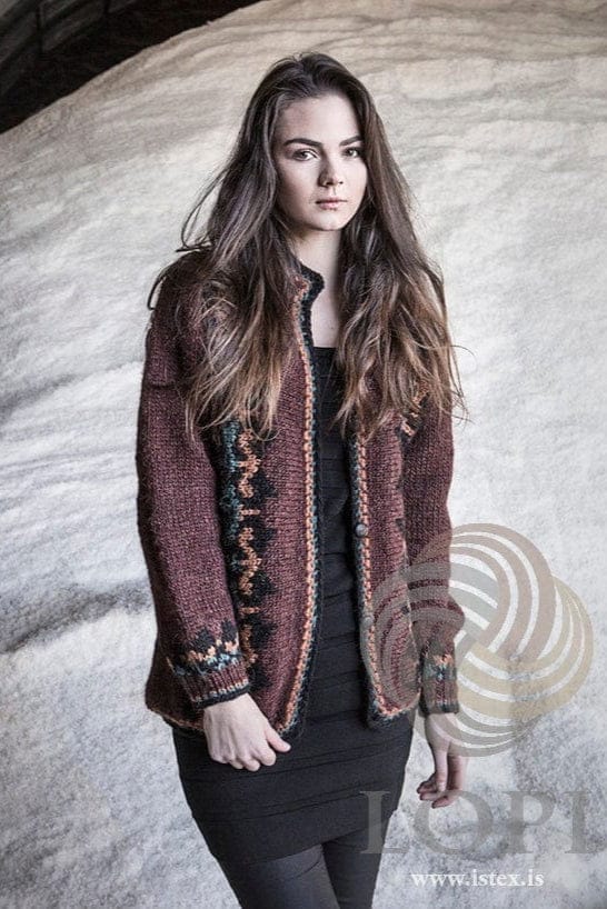 The auburn Lilja worn open, shown in the pattern’s own colours — sheep sorrel with black bands of teal and amber motifs down both fronts and near the cuffs, and a crocheted edge. The black top and dress and the black leggings are not part of the pattern