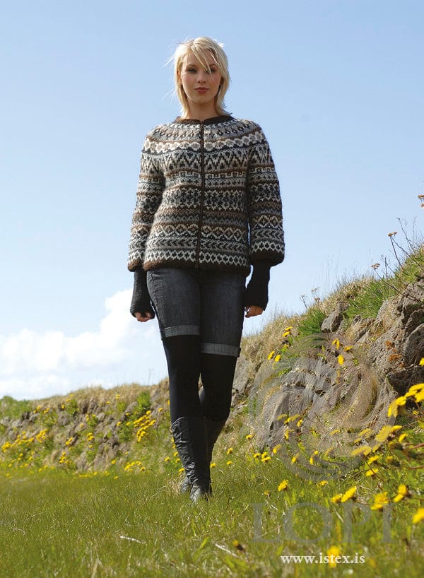 Young woman with short blonde hair walking on grass below a grassy bank in Saga 2, shown in the pattern’s own colours — a Léttlopi zip cardigan patterned all over in bands of small motifs in chocolate, white, black heather, greys, beige and oatmeal, with a chocolate neckband. The long dark arm warmers, the rolled-up denim shorts, the black tights and the black boots are not part of the pattern; the zipper is not included