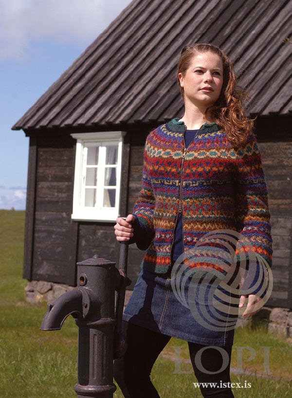 Young woman with long curly auburn hair by a black wooden house with a white window, one hand on an old iron water pump, in Saga 1, shown in the pattern’s original colours (orange, mustard, camel and hazel are no longer made) — a Léttlopi zip cardigan patterned all over in bands of small motifs in reds, orange, yellow, greens, teal, blues, purples and browns, with a dark green neckband. The navy top under it, the denim skirt and the black tights are not part of the pattern; the zipper is not included