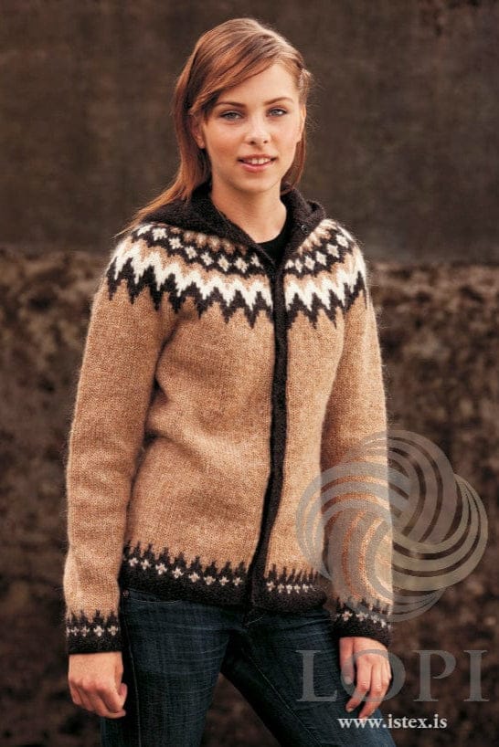 Young woman with long auburn hair against a dark background, wearing the brown Var in the pattern’s own colours — a barley cardigan with a yoke of black-sheep and white zigzags and small white diamonds, black-sheep front bands and border, and the hood lying round her neck. The black top under it and the dark blue jeans are not part of the pattern