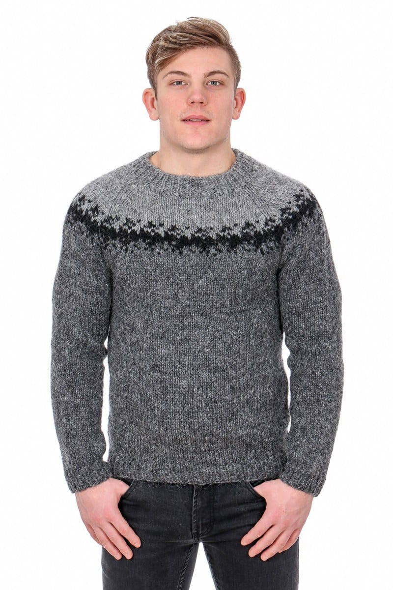 Sátt knitted in greys — a dark grey heather sweater with a black jagged band and a lighter grey upper yoke with raglan lines — on a young man against a white background. This is the photo of our dark grey kit, in our own colours, not a version in the pattern. The dark grey jeans are not part of the pattern