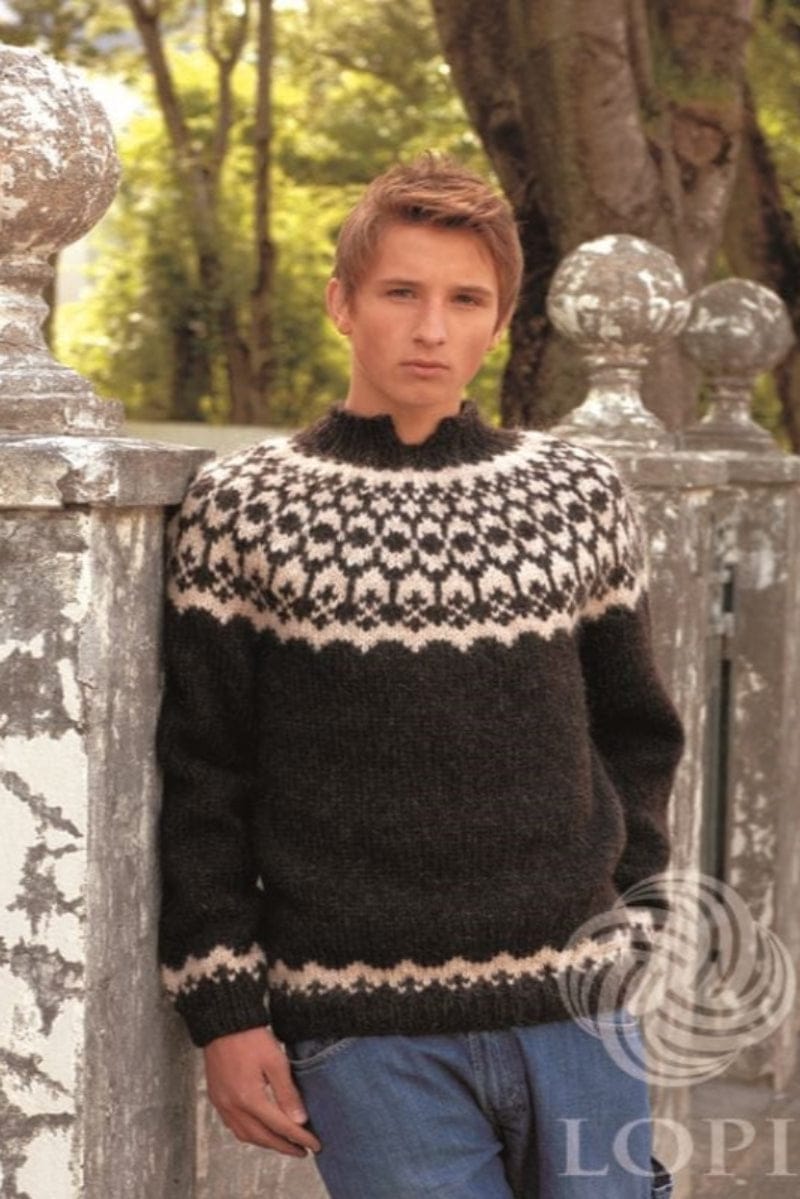 Young man with short reddish-brown hair leaning on an old stone gate pillar with flaking white paint, stone balls on the pillars and trees behind, wearing Virki in the pattern’s black version — sheep black with an ecru heather yoke of small crosses, arches and round dots above a wavy edge, a narrow ecru border above the hem and cuff rib, and a black ribbed collar. The jeans are not part of the pattern; a LOPI logo is printed in the corner
