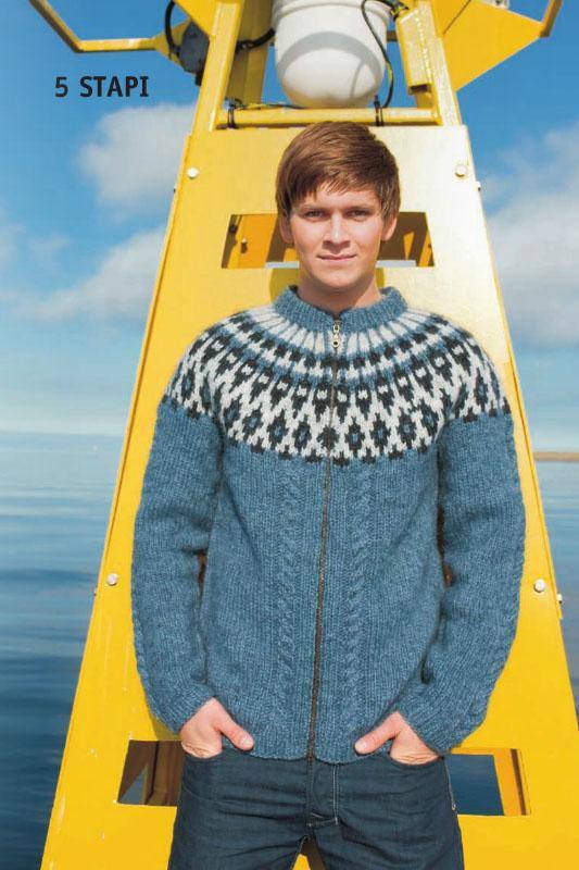 Young man with brown hair, hands in his pockets, in front of a yellow metal structure with sky and sea behind, wearing the blue Stapi in the pattern’s own colours — a light indigo cardigan zipped up the front, with cables beside the zip and a yoke of black and ash stars and diamonds. The jeans are not part of the pattern and the zip is not included. “5 STAPI” is printed on the photo