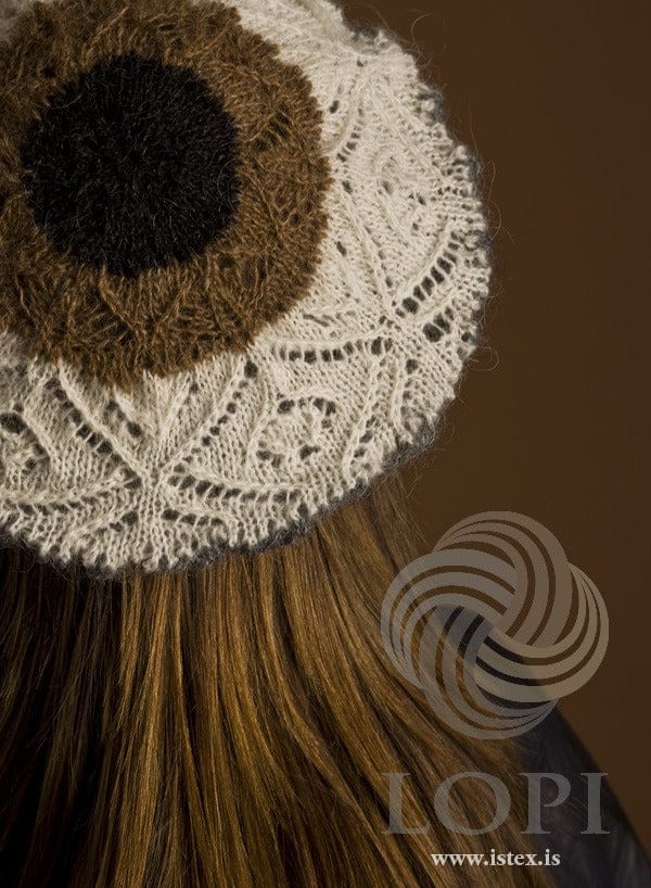 The top of the Mark beret from behind, in the pattern’s own colours — a black heather centre, a ring of tawny, a wide ring of white lace with large eyelet diamonds, and dark grey at the rim. The dark top at her shoulder is not part of the pattern