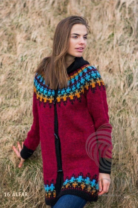 Woman in front of dry grass in Álfar, shown in the pattern’s own colours — a long garnet red Léttlopi cardigan with a round yoke and a border above the black rib hem in orange, golden, turquoise glacier blue and light heaven blue with black; black front bands, dark toggles, and the black hood lying down behind the neck. The blue jeans are not part of the pattern