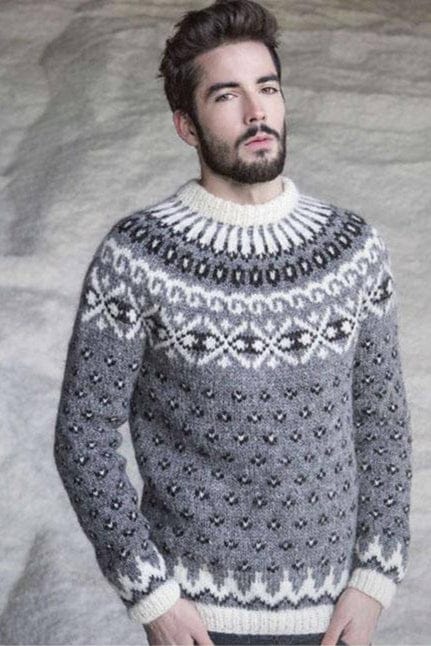 Bearded man against a pale, rough-textured background in Gnótt, shown in the pattern’s own colours — a grey heather Plötulopi sweater with small black-and-white lice all over, white pointed shapes above the white rib at the hem and cuffs, and a round yoke of a black band with white diamonds, white scrolls, a row of black shapes and white stripes up to a white rib collar. The dark trousers are not part of the pattern