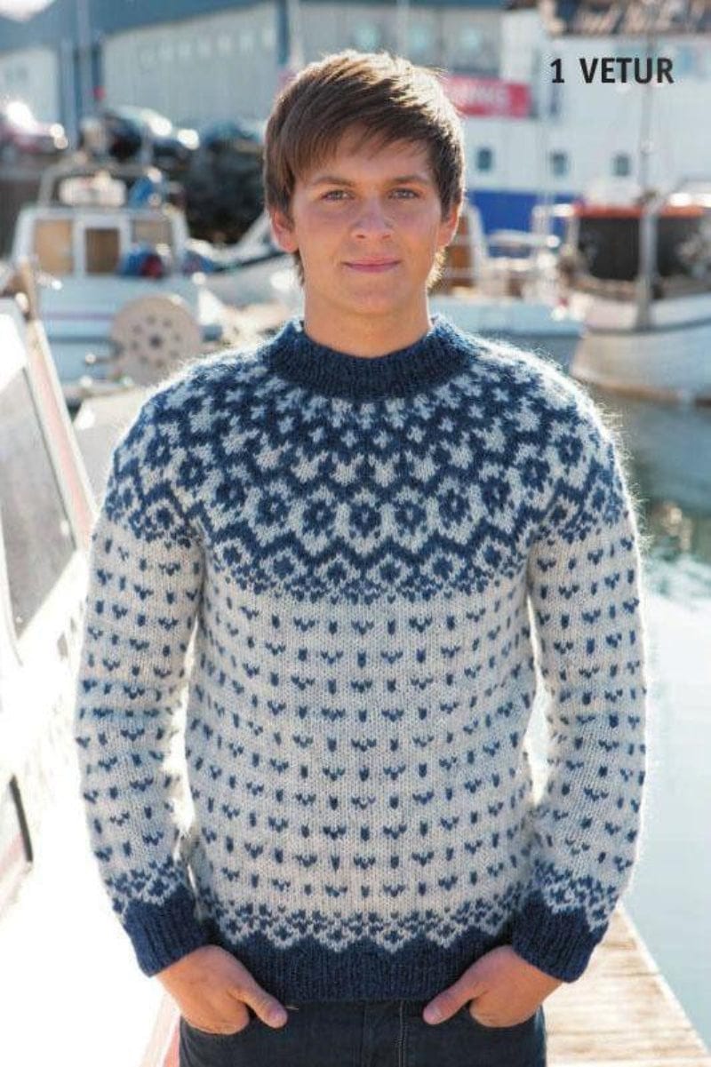 Young man with brown hair on a quay with boats behind, wearing the grey and blue Vetur in the pattern’s own colours — ash grey with small winter blue motifs, winter blue rib and neck, and a winter blue patterned yoke. The words “1 VETUR” are printed in the corner of the photo. The dark jeans are not part of the pattern