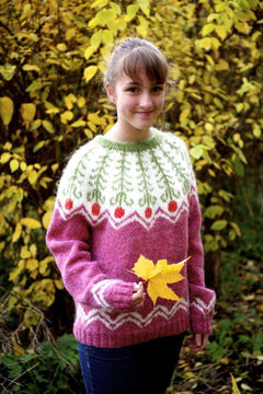 Girl among yellow autumn leaves in the Fern Flower sweater — pink heather with a white yoke of green ferns above red fern flowers, white zigzags above the hem and on the cuffs, and a green collar (shown in the original colours — this kit has crimson red for the light red flowers)