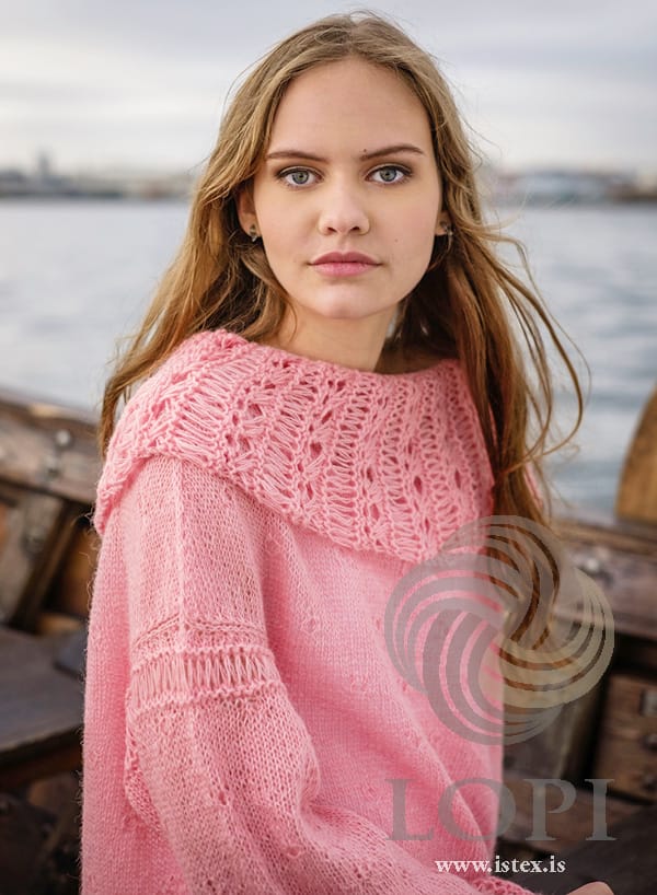 Close-up of a woman on a wooden boat in Askja, in the pattern’s original colour, blush — the deep collar of eyelets and long stitches standing out round the shoulders, and the band of long stitches round the top of the sleeve. Her earrings are not part of the pattern