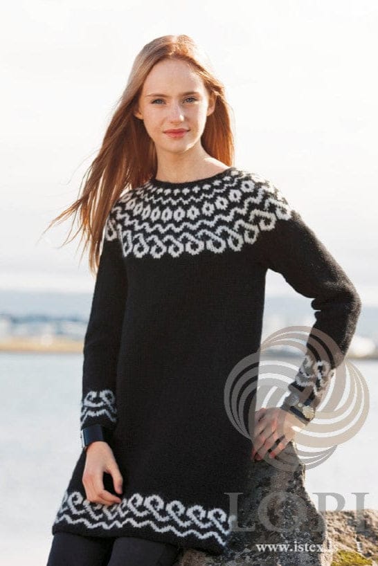Young woman with long red-blonde hair blowing in the wind, leaning on a rock by the water, wearing the short Keðja in the pattern’s black and white — a black dress with a white yoke of dots, small diamonds, waves and linked loops, and the same white border above the hem and the cuffs. The dark leggings, the wide black cuff bracelet and the leather cuff with silver ornaments are not part of the pattern