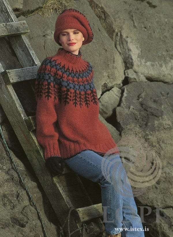 Woman sitting against a wooden ladder on grey rocks in the Lopi 12 no. 1 sweater and beret — a brick-red body, a yoke of black fir trees and blue and black checks, deep ribbed hem and cuffs, a ribbed stand-up collar, and black and blue blanket stitch along the edges (shown in the original colours — this kit has amber heather, more orange, for the rust 9392, and denim heather for the steel blue 9300). The jeans, black gloves and earrings are not part of the pattern