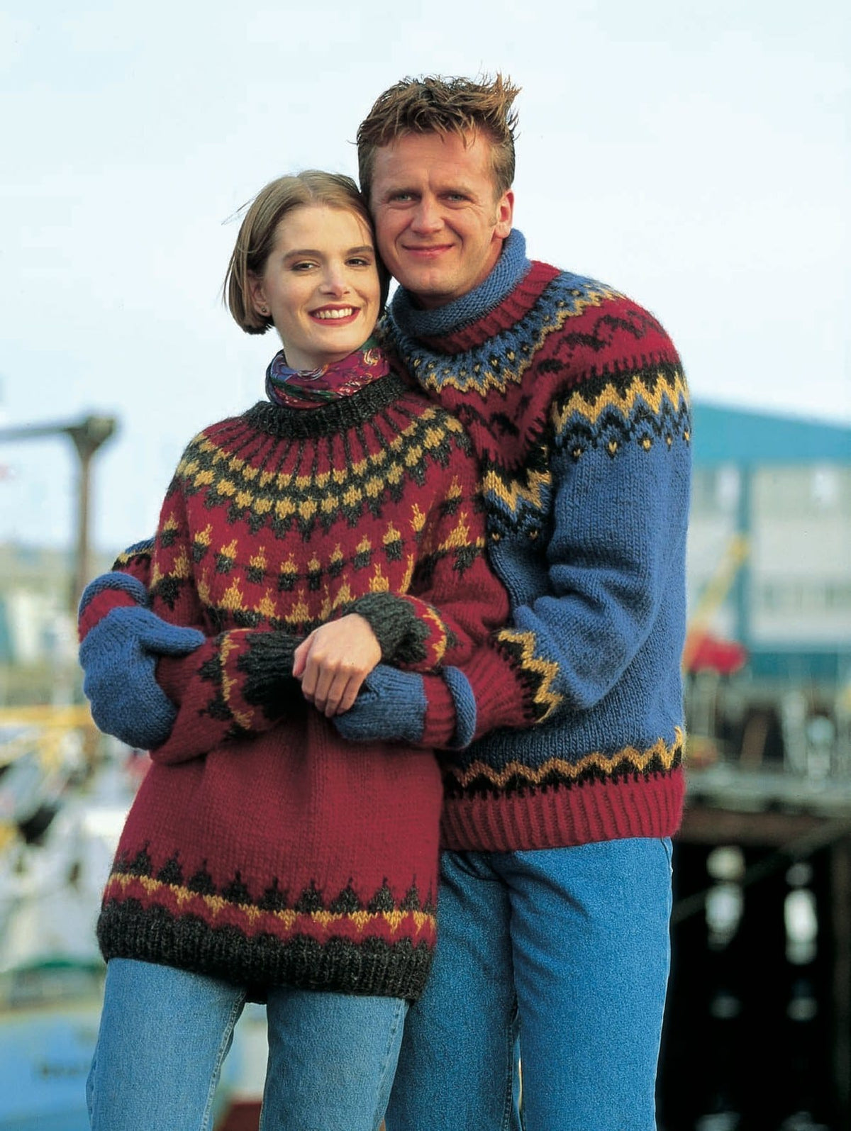 Couple by a harbour. The man (right) wears the Lopi 12 no. 13 sweater — mid blue with red rib at the hem, a yoke of gold and black zigzags with a red band, and a blue roll collar over red rib — and the woman wears its blue mittens with red ribbed cuffs (shown in the original colours — this kit has denim heather, happy red and a deeper golden heather for 0098, 9389 and 9332). Her red sweater is Lopi 12 no. 11, a different pattern; the jeans and scarf are not part of it