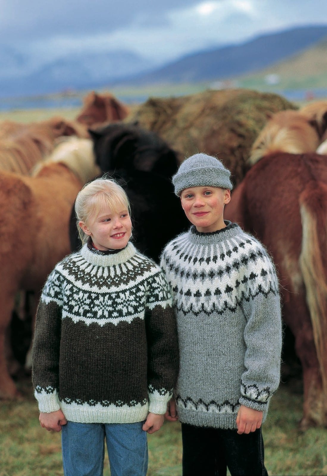 Two children in front of Icelandic horses. The boy on the right wears the Lopi 12 no. 27 sweater — light grey ash heather with a white and black heather yoke: a scalloped white band, small dark diamonds and white stems at the neck, with a matching border above the hem and cuffs — and the plain grey rolled-brim hat from the same leaflet, in the pattern’s own colours. His dark trousers are not part of the pattern; the child on the left wears Lopi 12 no. 29, a different pattern, with jeans and a neck scarf