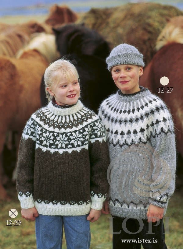 Two children in front of Icelandic horses. The girl on the left wears No. 29, the brown version of this pattern, in its own colours — chocolate brown with white rib and a folded white neckband, small motifs above the rib, and a yoke with black sheep heather star motifs on a white band and white rays up to the neck. Her bandana and jeans are not part of the pattern; the boy on the right wears Lopi 12 no. 27, a different pattern, with a hat and dark trousers
