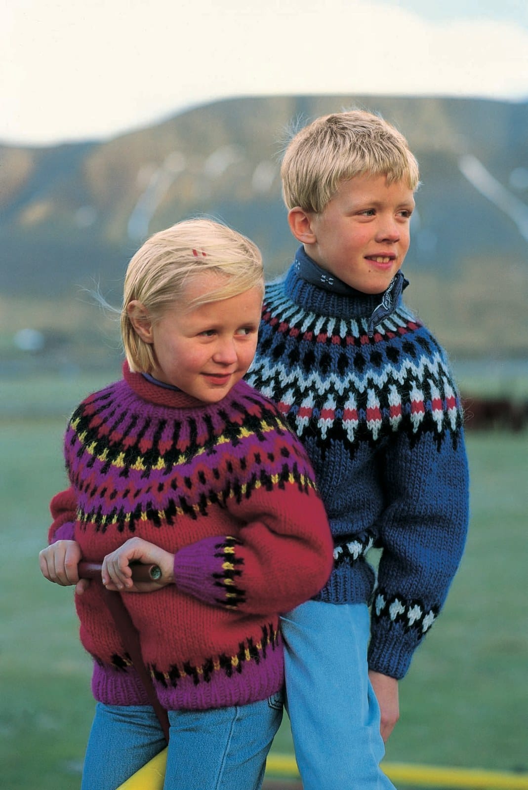 Girl in front in the Lopi 12 no. 30 sweater — a scarlet red body, purple rib at the hem and cuffs with a purple, black and gold border above, a yoke of purple bands, black and gold rows and purple and black rays, and a red roll collar, shown in the pattern’s original colours (0078, 9010 and 9213 are no longer made). Her hair clip, the top under her collar, the handle she holds and her jeans are not part of the pattern; the boy behind her wears Lopi 12 no. 31, a different pattern