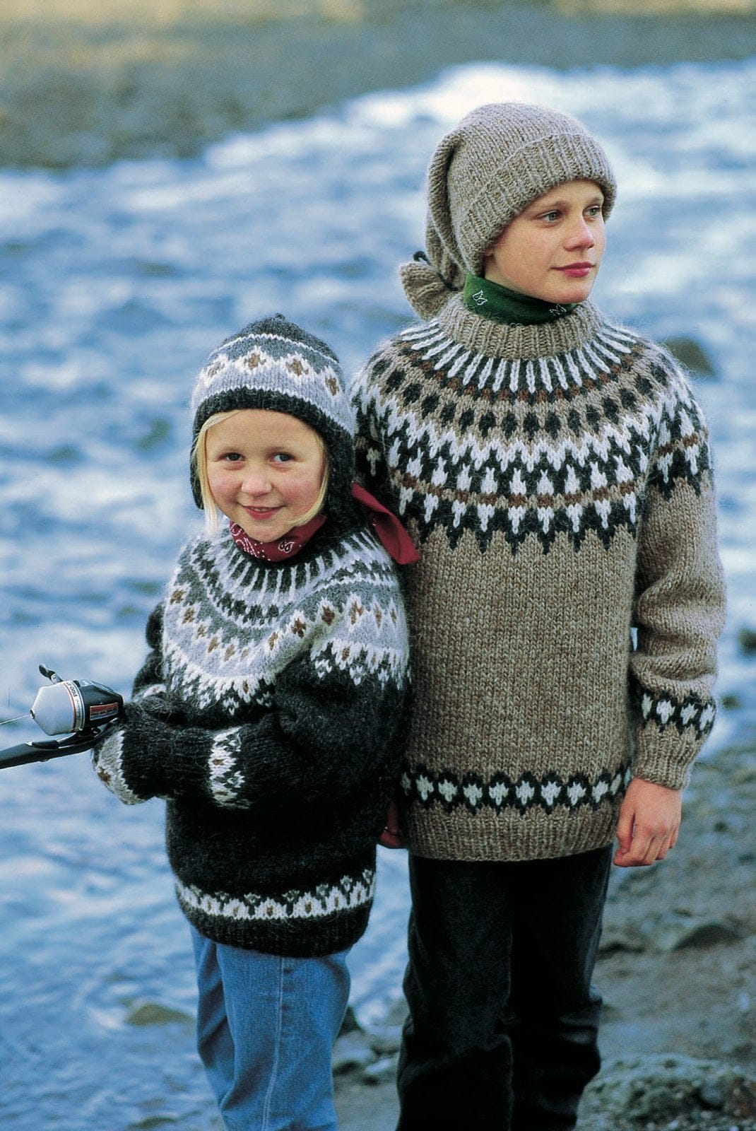 By the sea: the girl on the left wears the Lopi 12 no. 34 set — a charcoal sweater with a yoke of white, light grey, dark grey and acorn brown and a white border above the hem, the matching ear-flap hat and the mittens. Her red scarf, the jeans and the fishing rod are not part of the pattern; the boy on the right wears Lopi 12 no. 32, a different pattern, sold as its own kit